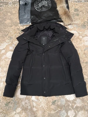 Canada Goose wyndham - Snygg svart dunjacka från Canada Goose med avtagbar pälskrage och hög krage. Jackan har dragkedja och knappar framtill, två stora fickor och diskret logotyp på ärmen. Tillverkad i slitstarkt material och fodrad med dun för extra värme. 