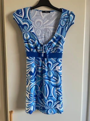 Blåvit mönstrad topp  - Säljer en blå och vit topp från Jane Norman med grafiskt mönster, djup v-ringning och bred midjerem med knyt i ryggen. Toppen har korta ärmar och är tillverkad i ett mjukt, stretchigt material som känns skönt mot huden. Perfekt för dig som gillar färg och unika mönster!  Är i storlek s/m.