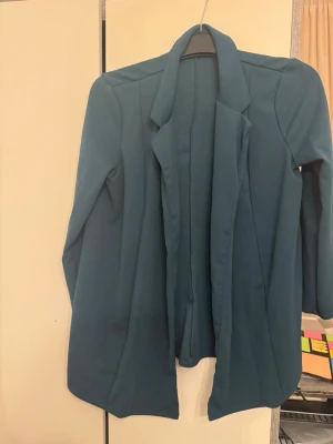 Mörkgrön kostym med kavaj och byxor - Snygg mörkgrön kostym med klassisk kavaj och matchande byxor. Kavajen har en rak passform och långa ärmar, medan byxorna har pressveck och hög midja. Perfekt för dig som vill ha en stilren och modern look.