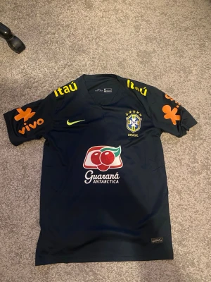 Brasilien träningströja Nike svart - Snygg svart träningströja från Nike med Brasilien CBF-logga på bröstet, gula och orangea sponsortryck samt Guaraná Antarctica-logga framtill. Kortärmad modell i lätt och ventilerande material, perfekt för fotbollsträning.