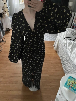 Svart långklänning med blommigt mönster - Trendig svart långklänning med små gula blommor, v-ringad och med puffiga långärmar. Klänningen har knappar framtill och markerad midja för en snygg siluett. Perfekt för dig som vill sticka ut med en romantisk look.