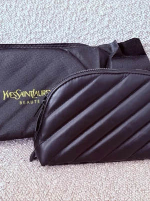 Svart necessär från Yves Saint Laurent, sminkväska  - Säljer en svart necessär från Yves Saint Laurent med gul logotyp och dragkedja. Materialet är slitstark nylon och väskan har en enkel, avlång form. Perfekt för att förvara smink eller småsaker. Medföljer även en mindre quiltad svart väska med dragkedja. 20*13*5cm