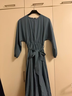 Blå långklänning med knytband - Långklänning i mjukt blått tyg med trekvartsärm och rund halsringning. Klänningen har ett brett knytband i midjan för en markerad siluett och stängs med en knapp i nacken. Perfekt för dig som gillar enkel men stilren design.