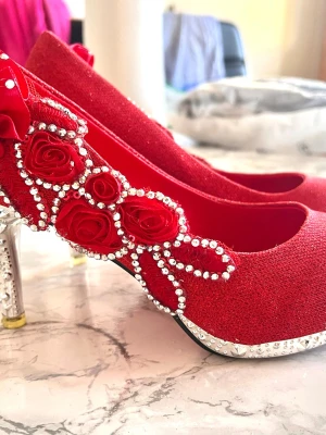 Glittrande Röda pumps med pärlor och rosor - Unika röda pumps med glittrande yta, dekorerade med röda tygrosor och pärlor längs sidan. Klacken är silverfärgad med detaljer och har en elegant form. Perfekta för dig som vill sticka ut och älskar detaljerade skor.