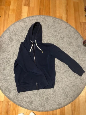 Marinblå hoodie från Polo Ralph Lauren - Snygg marinblå hoodie från Polo Ralph Lauren med dragkedja, vit dragsko och klassisk gul broderad logga på bröstet. Hoodien har kängurufickor och ribbade muddar. Perfekt för en avslappnad och stilren look.
