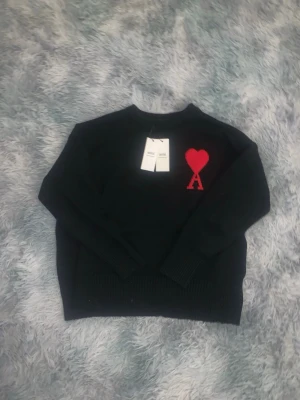 Svart stickad tröja Ami Paris - Svart stickad tröja från Ami Paris med röd hjärtlogga och bokstaven A på bröstet. Tröjan har rund halsringning, långa ärmar och ribbade muddar vid ärmslut och nederkant. Perfekt för dig som gillar stilrena plagg med en twist.