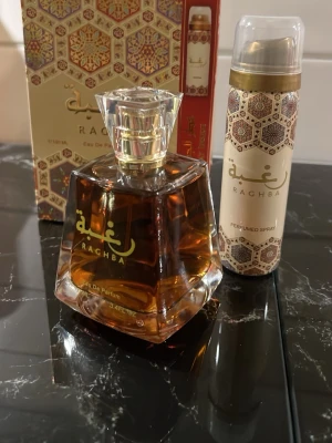 Raghba Eau De Parfum 100ml - Raghba Eau De Parfum 100ml med elegant fyrkantig glasflaska och guldiga detaljer. Doften har en orientalisk känsla och flaskan har en lyxig, genomskinlig design med bärnstensfärgad vätska. Medföljer även en matchande perfumed spray.