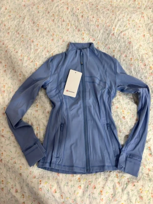 Blå define träningsjacka från Lululemon - Snygg ljusblå träningsjacka från Lululemon med hel dragkedja framtill, hög krage och två fickor med dragkedja. Tillverkad i ett mjukt och stretchigt funktionsmaterial som andas. Perfekt för träning eller som ett extra lager på väg till gymmet. Storlek 4 men skulle säga att den är som en S