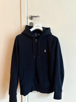 Mörkblå hoodie från Polo Ralph Lauren - Polo Ralph Lauren navy kofta, köpt på NK i Göteborg, kvitto finns och medföljer. Storlek M. Perfekt för en avslappnad och stilren look.           (litet hål i knappt synligt)