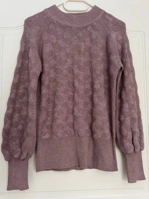Helt ny ljuslila/smutsrosa stickad tröja med struktur  - Supersnygg ljuslila stickad tröja med unik strukturstickning och ballongärmar. Tröjan har bred ribbad mudd i nederkant och vid ärmslut samt en högre rund halsringning. Perfekt för dig som vill ha något extra till din outfit. Helt ny! 