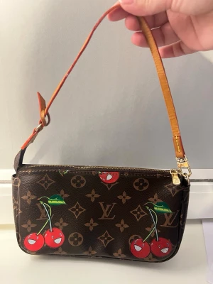 Louis Vuitton Monogram Cherry Pochette vintage - Snygg brun handväska från Louis Vuitton med klassiskt monogrammönster och lekfulla körsbärsmotiv. Väskan har guldfärgade detaljer och justerbart axelband i ljust läder. Perfekt för dig som vill sticka ut med en unik accessoar.