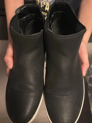 Svarta Chelsea boots i skinn - Säljer ett par klassiska svarta Chelsea boots i slätt skinn med elastiska paneler på sidorna och vit sula. Bootsens minimalistiska design gör dem enkla att matcha med olika outfits. Perfekta för dig som gillar stilrena och tidlösa skor.