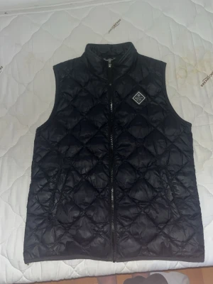 Svart quiltad dunväst med dragkedja - Snygg svart dunväst. Västen har hel dragkedja framtill och en liten patch på bröstet. Perfekt att slänga över en hoodie eller tröja för extra värme. Materialet är glansigt och lätt, vilket gör den enkel att bära. Säljer den för den slutat passa mig 