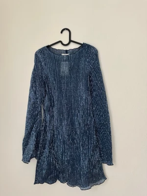 Blå glittrig klänning med paljetter - Supersnygg blå klänning täckt av glittriga paljetter. Klänningen har lång ärm, rund halsringning och lätt utställd nederkant. Aldrig använd 