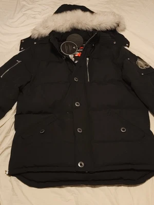 Svart Moose Knuckles med vit päls och silver detaljer  - Svart pufferjacka från Moose Knuckles med avtagbar huva och vit päls. Jackan har flera fickor med dragkedja och knappar, samt logga på ärmen. Perfekt för kalla vinterdagar och riktigt snygg till streetwear. Pris kan diskuteras. XXL passar som XL