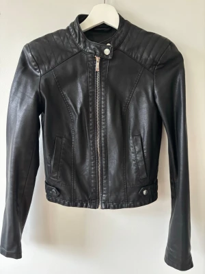 Svart skinnjacka från Amisu - Snygg svart skinnjacka från Amisu med quiltade axlar och ståkrage. Jackan har dragkedja framtill, två fickor och dekorativa knappar vid midjan. Insidan har ett blommigt foder som ger en cool kontrast. Perfekt för dig som vill ha en stilren och trendig look.