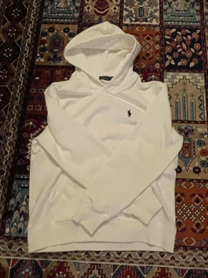 Vit hoodie från Polo Ralph Lauren - Snygg vit hoodie från Polo Ralph Lauren med klassisk broderad logga på bröstet. Hoodien har huva, känguruficka och ribbade muddar. Perfekt för en clean och stilren look. Materialet känns mjukt och bekvämt mot huden.
