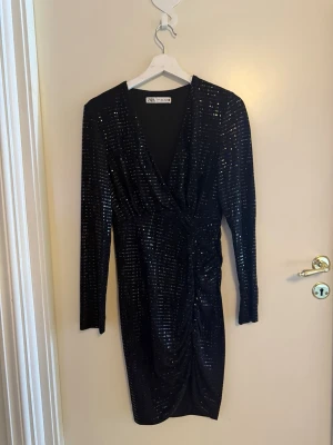 Svart glittrig omlottklänning Zara - Snygg svart omlottklänning från Zara med glittriga detaljer. Klänningen har lång ärm, v-ringning och rynkad effekt i midjan. Perfekt för dig som vill sticka ut med en elegant look.