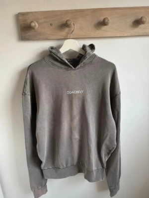 Grå hoodie från Coach Ivy - Säljer en grå hoodie från Coach Ivy med broderad vit logga på bröstet. Hoodien har en klassisk passform med huva och ribbade muddar vid ärmslut och nederkant. Tillverkad i mjukt bomullsmaterial som är perfekt för en avslappnad stil.