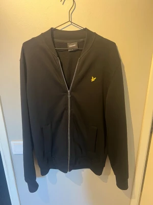 Svart zip hoodie från Lyle & Scott - Snygg svart hoodie från Lyle & Scott med dragkedja framtill och två fickor. Klassisk gul logga på bröstet och ribbade muddar vid ärmar och nederkant. Tillverkad i mjukt material som känns skönt mot huden. Perfekt för en avslappnad och stilren look. Nypris:799kr! Kanppt använd!