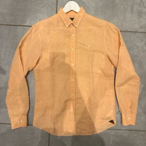 Linneskjorta från Morris - Snygg persikofärgad skjorta i linne med klassisk krage och långa ärmar. Skjortan har knäppning framtill, manschetter med knappar och en diskret broderad logga på bröstet. Perfekt för en avslappnad men stilren look. 100% Linne