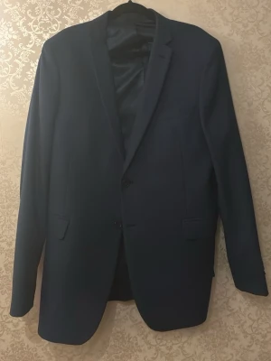 Mörkblå klassisk kavaj - Snygg mörkblå kavaj med två knappar framtill och klassiska fickor. Kavajen har slimmad passform och är helfodrad med slätt innerfoder. Materialet ser ut att vara en blandning av polyester och bomull, perfekt för en stilren look. Passar dig som vill ha en tidlös och elegant stil.