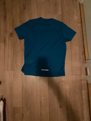 Blå Nike tränings-tshirt - Snygg blå tränings-tshirt från Nike med vit logga på bröstet och tunna vita detaljer vid axlarna. Tillverkad i ett lätt och snabbtorkande material som passar perfekt till gymmet eller löprundan. Klassisk passform med rund hals och korta ärmar.