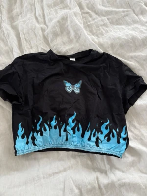 Svart croppad topp med blå fjäril - Cool svart croppad topp med blå fjäril på framsidan och blåa flames längs nederkanten. Kortärmad modell , perfekt till jeans eller kjol för en edgy look.