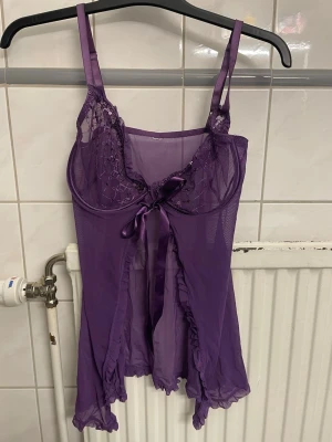 Lila mesh body med spetsdetaljer - Säljer en lila body i transparent mesh med tunna axelband och dekorativa spetsdetaljer upptill. Framtill finns en söt rosett och volangkant längs nederdelen. Rosetten är ett sidenband man kan knyta själv. Perfekt för dig som gillar feminina underkläder.