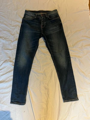 Mörkblå jeans från Nudie Jeans - Säljer ett par mörkblå jeans från Nudie Jeans med klassisk femficksmodell och knappgylf. Jeansen har en snygg tvätt med slitningar på låren och raka ben. Perfekta för dig som gillar en tidlös och avslappnad stil.