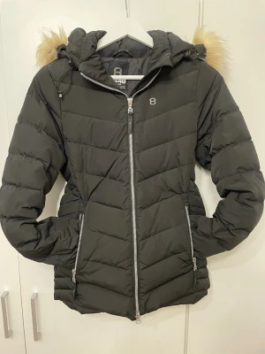 Svart dunjacka med fuskpäls från 8848 - Snygg svart dunjacka från 8848 med avtagbar huva och beige fuskpäls. Jackan har två dragkedjeförsedda fickor, silverfärgade detaljer och quiltad design. Perfekt för kalla vinterdagar och riktigt varm.