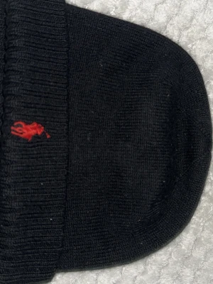 Svart mössa från Polo Ralph Lauren - Säljer en svart ribbstickad mössa från Polo Ralph Lauren i mjuk merinoull. Mössan har en klassisk design med uppvikt kant och den ikoniska röda Polo-loggan broderad framtill. Perfekt för att hålla värmen och ge en stilren touch till din outfit.