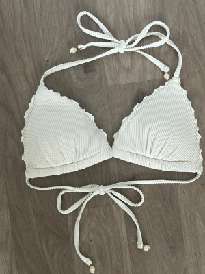 Vit ribbad trekantsbikini - Snygg vit trekantsbikini med ribbad struktur och knytband i både nacke och rygg. Bikinin har vågiga kanter och små pärldetaljer på snörena för en extra touch. Perfekt för lata dagar på stranden eller poolen!