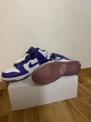 Nike Dunk Low lila och vita sneakers - Säljer ett par Nike Dunk Low sneakers i lila och vitt med klassisk Swoosh på sidan. Skorna har en låg siluett, vita paneler i läder och lila detaljer samt genomskinlig rosa sula. Perfekta för dig som vill sticka ut med färg på fötterna.