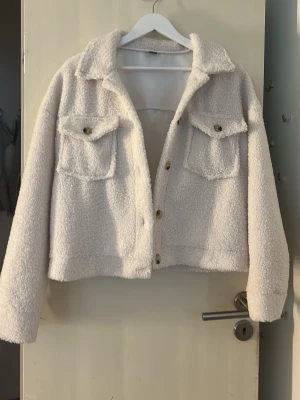 Vit cropped teddyjacka med fickor - Säljer en vit teddyjacka i croppad modell med två bröstfickor och stora guldfärgade knappar. Jackan har bred krage och är gjord i mjukt, fluffigt teddy-material som ger en riktigt cozy vibe. Perfekt för dig som vill ha en trendig och bekväm jacka till hösten eller vintern.