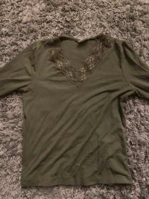 Olivgrön topp med spets från Gina Tricot - Snygg olivgrön topp från Gina Tricot med v-ringning och dekorativ spetsdetalj runt halsen. Toppen har långa ärmar och är tillverkad i mjukt material som känns skönt mot huden. Perfekt för dig som vill ha en enkel men ändå stilren look.