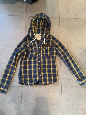 Rutig overshirt med huva från Hollister - Snygg overshirt från Hollister i blått och gult rutigt mönster. Modellen har huva med vita dragsnören, två bröstfickor med knapp och broderad logga. Overshirten har långa ärmar och knäppning framtill. Perfekt för en avslappnad streetstil.