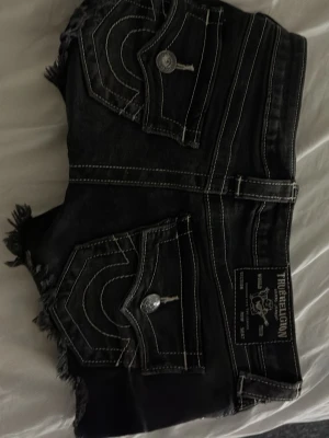 Svarta jeansshorts True Religion - Svarta jeansshorts från True Religion med slitna kanter och tydliga vita kontrastsömmar. Shortsen har stora bakfickor med lock och silverfärgade knappar samt en tydlig logotyp-patch bak. Klassisk femficksmodell i denim.