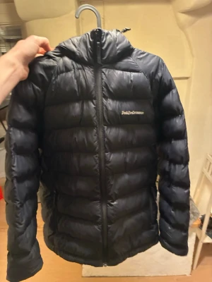 Svart dunjacka från Peak Performance - Säljer en svart dunjacka från Peak Performance med huva och dragkedja framtill. Jackan har en klassisk puffer-design med horisontella sömmar och logga på bröstet. Perfekt för kalla vinterdagar och riktigt skön att bära.