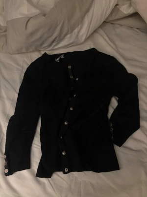 Svart kofta med knappar från Gina Tricot - Svart långärmad kofta från Gina Tricot med silvriga knappar längs hela framsidan och vid ärmsluten. Smal passform och rund halsringning. Perfekt att styla med jeans eller kjol för en snygg look.