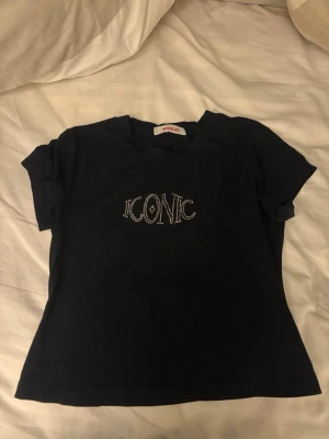 Svart t-shirt med strassdetalj - Svart t-shirt från Wishful med korta ärmar och texten 'ICONIC' i strass på bröstet. T-shirten har en klassisk passform och är gjord i mjuk bomull, perfekt för en trendig och avslappnad look.