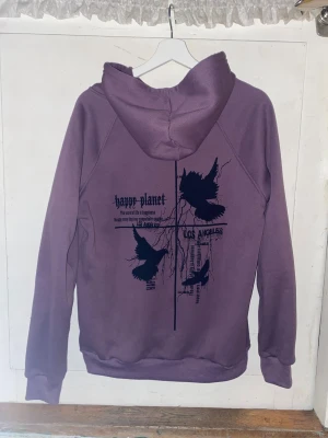 Lila hoodie med tryck - Lila hoodie med svart tryck på både fram- och baksida. Framsidan har texten 'happy planet' och baksidan har grafiskt tryck med fåglar och text. Hoodien har huva med svarta snören och en stor magficka. Perfekt för en avslappnad streetstyle. Storlek S