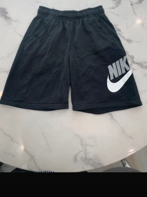 Svarta shorts från Nike med logga - Svarta shorts från Nike med stor vit logga och swoosh på vänster ben. De har elastisk midja och två sidofickor. Tillverkade i mjukt bomullsmaterial, perfekta för en avslappnad och sportig stil.