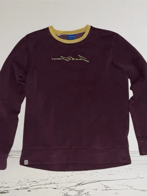 Jack & Jones tröja - Vinröd sweatshirt från Jack & Jones med gul kontrastfärgad krage och broderad logga på bröstet. Tröjan har rund halsringning och långa ärmar. Perfekt för en avslappnad och stilren look. Storlek S.
