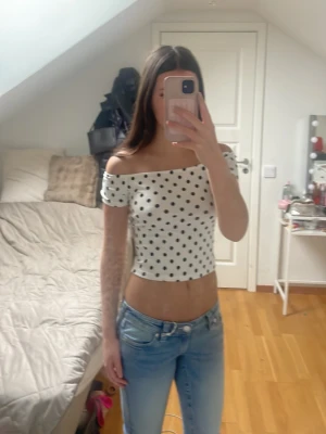 Vit offshoulder prickig topp - Säljer en vit offshoulder topp med svarta prickar. Toppens passform är tight och den är croppad, vilket ger en modern look. Perfekt för dig som gillar att sticka ut med ett klassiskt mönster. Materialet känns mjukt och stretchigt, troligen bomull eller polyester.