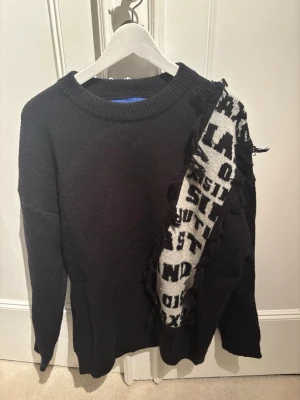 Svart stickad tröja Elfric Eden - Cool svart stickad tröja från Elfric Eden med en unik vit panel med svart text och fransdetaljer på ena sidan. Tröjan har rund halsringning och långa ärmar. 