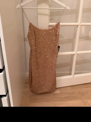 Champagnefärgad paljettklänning - Säljer en glittrig champagnefärgad kortklänning från HEY med tunna axelband och täckt av paljetter. Klänningen har en rak passform och är perfekt för dig som vill sticka ut. Materialet känns mjukt och följsamt mot huden.