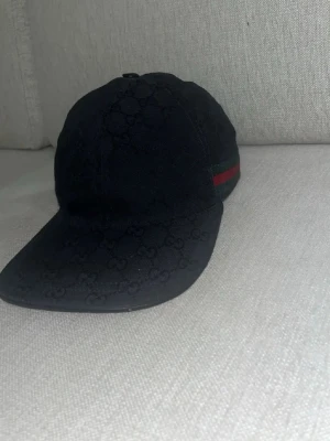 Svart keps från Gucci med logomönster - Snygg svart keps från Gucci med diskret GG-logomönster över hela kepsen och klassisk röd-grön rand på sidan. Kepsen har böjd skärm och justerbar passform. Perfekt för dig som vill ha en stilren accessoar med lyxig känsla.                              Pris kan sänkas vid snabbäffar!! Kvitto och dustbag ingår!! Möts upp i Stockholm !!