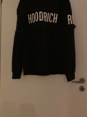 Svart hoodie från Hoodrich - Säljer en svart hoodie från Hoodrich med stor vit logga tryckt över bröstet och ryggen. Hoodien har huva och långa ärmar. Perfekt för dig som gillar streetwear och vill ha en stilren men ändå iögonfallande look.