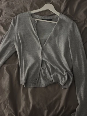 Grå basic kofta från H&M - En enkel och stilren grå kofta från H&M med rund halsringning och knäppning framtill. Koftan har långa ärmar och ribbade muddar vid ärmslut och nederkant. Perfekt att slänga över en topp för en clean look.